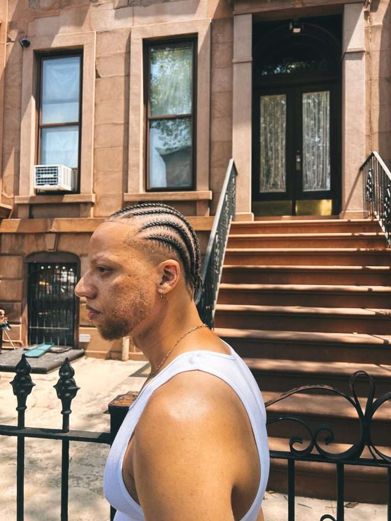 Najee Omar in profile with braided hair, standing at the iron fence of a Brooklyn brownstone stoop