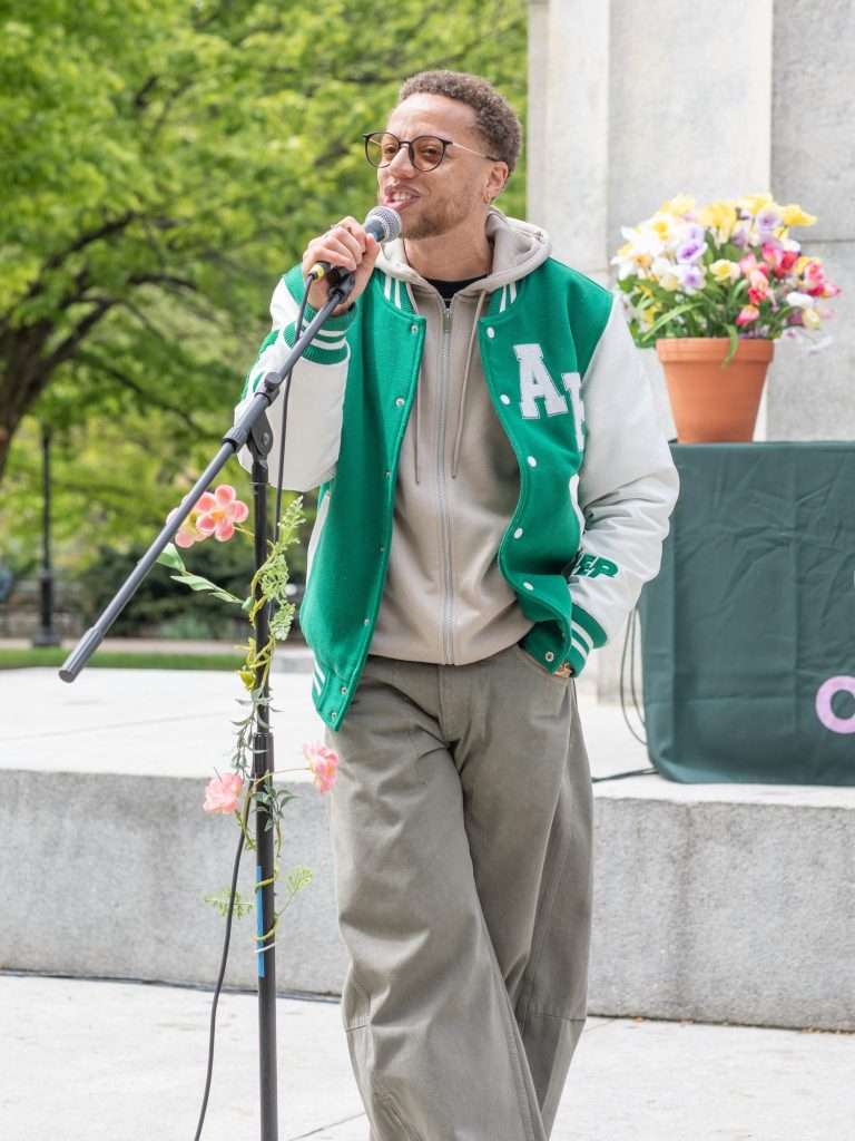 Najee Omar reading into a microphone outdoors at Poetry in the Park, in a green varsity jacket